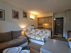 Een functionele leefruimte met een bank, een tafeltje, een kitchenette en een deur naar een andere kamer.