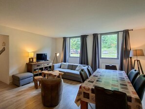 Sala de estar com sofá, banqueta, mesa de centro e televisão. Há janelas com cortinas, um abajur e vista para o exterior.