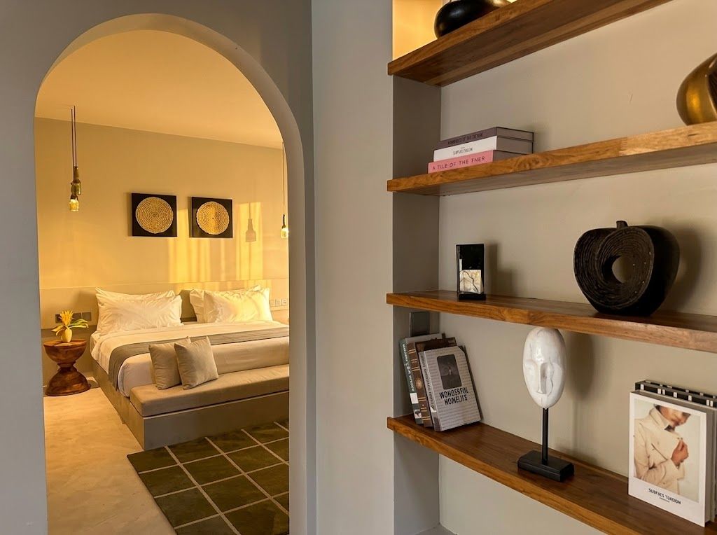 A bedroom with a bed, a bedside table with a lamp, and a shelf with books and decorative items.