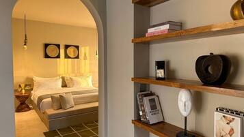 A bedroom with a bed, a bedside table with a lamp, and a shelf with books and decorative items.