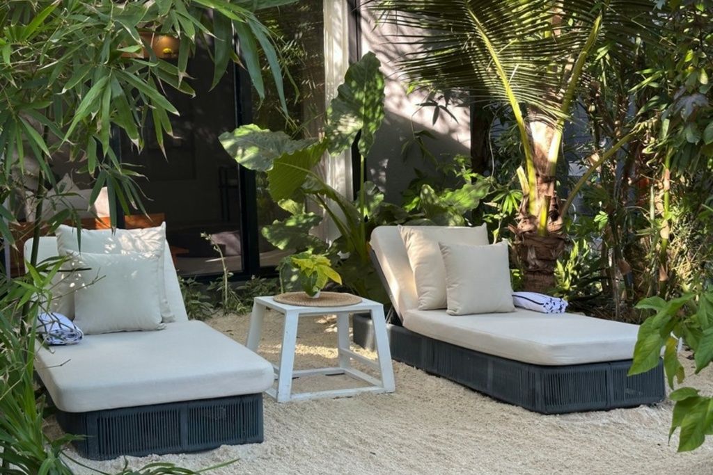 Un jardín con dos zonas de estar blancas y acolchadas, rodeado de abundante vegetación y una pequeña mesa blanca.