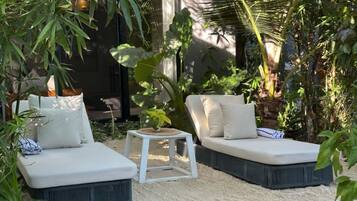 Un jardín con dos zonas de estar blancas y acolchadas, rodeado de abundante vegetación y una pequeña mesa blanca.