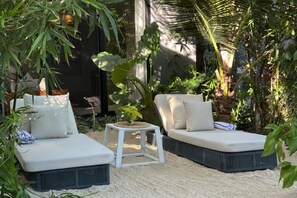 Un jardí amb dues zones d'seure amb seients blancs i coixins, envoltat de vegetació abundant i una taula petita blanca.