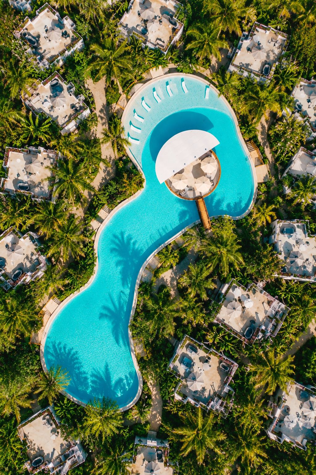 Vista aérea de un complejo turístico con una piscina serpenteante grande, rodeado de palmeras y edificios del complejo.