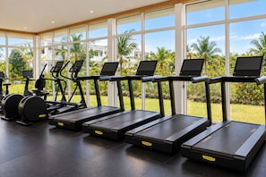 A gym with treadmills, stationary bikes, and ellipticals, large windows, and a view of palm trees and a building. - Maya Resort Zanzibar (Dongwe)