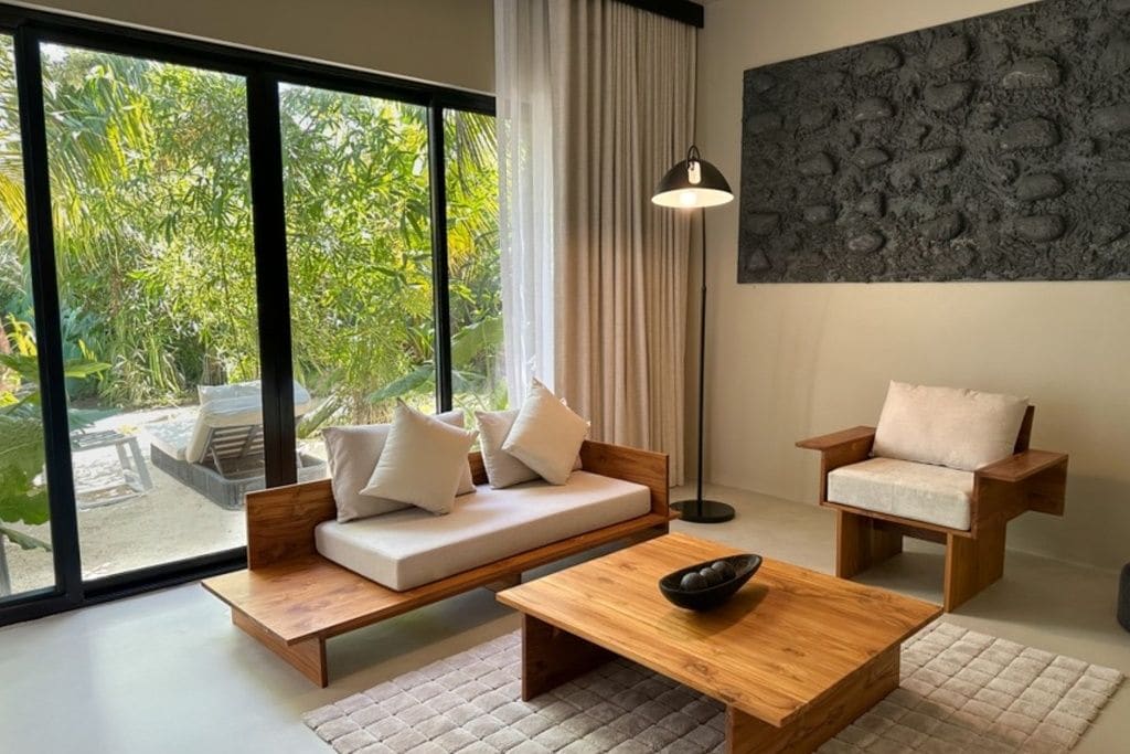 Un salon moderne comprenant un canapé, un fauteuil et une table basse, de grandes fenêtres offrant une vue sur la verdure, et une œuvre d’art murale à la texture intéressante.