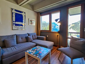 A living room with a sofa, a coffee table with a colorful print, a lamp, and a window with a view of mountains. - Renovated apartment between slopes and shops - 6 guests (Flaine)