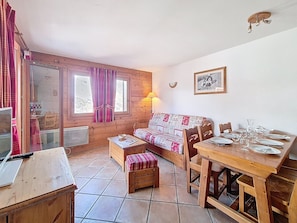 Ein gemütliches Zimmer mit Holzboden, einer Couch mit gemustertem Bezug, einem Esstisch mit Geschirr und Gläsern sowie einer Lampe.