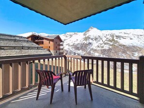 A balcony with two chairs and a table, overlooking a snow-covered mountain and a building with a sloped roof. - Spacious apartment near slopes - 6 guests (Les Menuires)