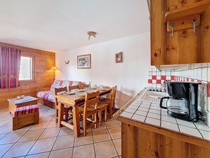 A cozy room with a wooden dining table, chairs, a bench, a small kitchen area with a coffee maker, and a window with curtains. - Spacious apartment near slopes - 6 guests (Les Menuires)