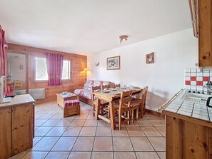 Ein gemütliches Blockhaus-Interieur mit Esstisch, Stühlen, einer Couch, einer kleinen Küchenzeile und einem Fenster mit Vorhängen.