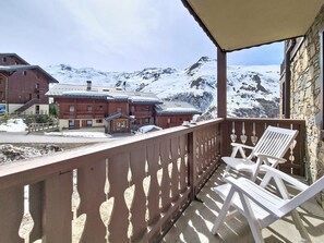 A balcony with white chairs, a view of snow-covered mountains, and a building with a red facade. - Spacious apartment near the slopes - 4 guests (Les Menuires)