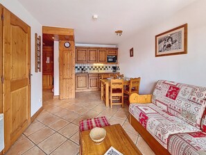A cozy living room with a sofa and a coffee table, a dining area with a table and chairs, and a kitchen with wooden cabinets and a tiled backsplash. - Spacious apartment near the slopes - 4 guests (Les Menuires)