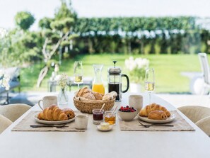 Ein Frühstückstisch gedeckt mit Croissants, einer Obstschale, Kaffee und Saft, mit Blick auf einen Garten und eine Rasenfläche.