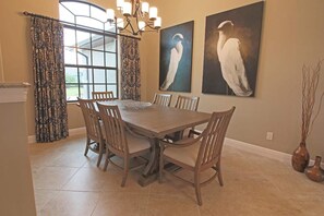 Een eetkamer met een grote tafel, stoelen, een kroonluchter en twee vogelschilderijen aan de muur.