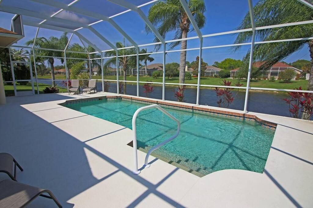 A covered swimming pool with clear water, surrounded by palm trees and houses in the background.