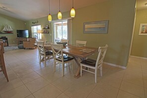 A dining area with a wooden table and white chairs, a television in the background, and a window with a view of the outdoors. - Lakeside Luxury #198 (Rotonda West)