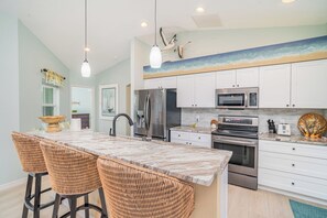 A modern kitchen with white cabinets, stainless steel appliances, a marble island with bar stools, and a coastal-themed wall mural. - This house is a 2 bedroom(s), 2 bathrooms, located in Rotonda West, FL. (Rotonda West)