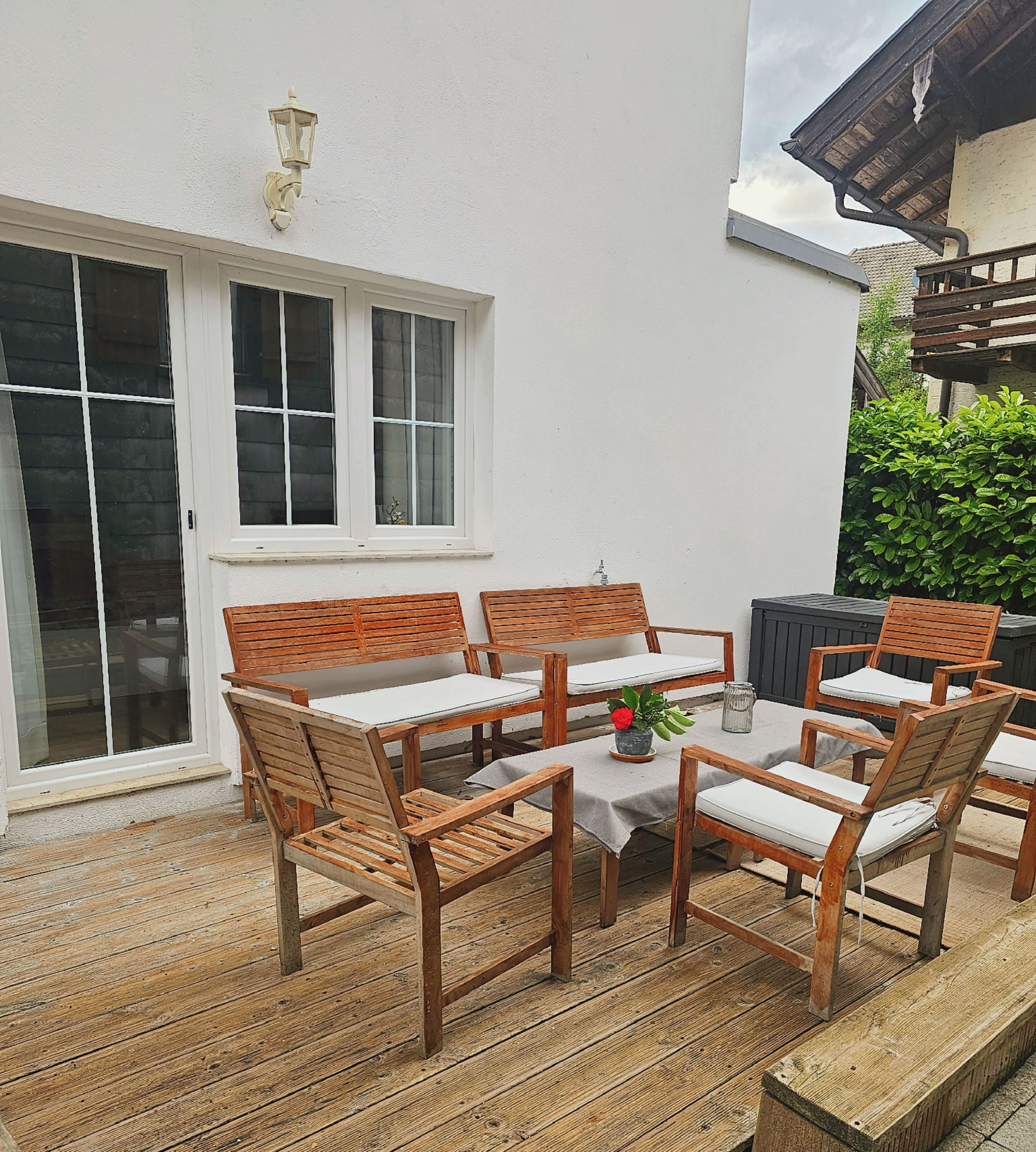 A wooden deck with a table and chairs, a white wall, and a window.
