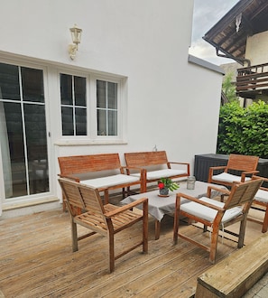 A wooden deck with a table and chairs, a white wall, and a window.