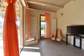 Ein Zimmer mit einer Schiebetür, einem Sessel, einem Fernseher auf einem Schrank und Blick auf Grünflächen.