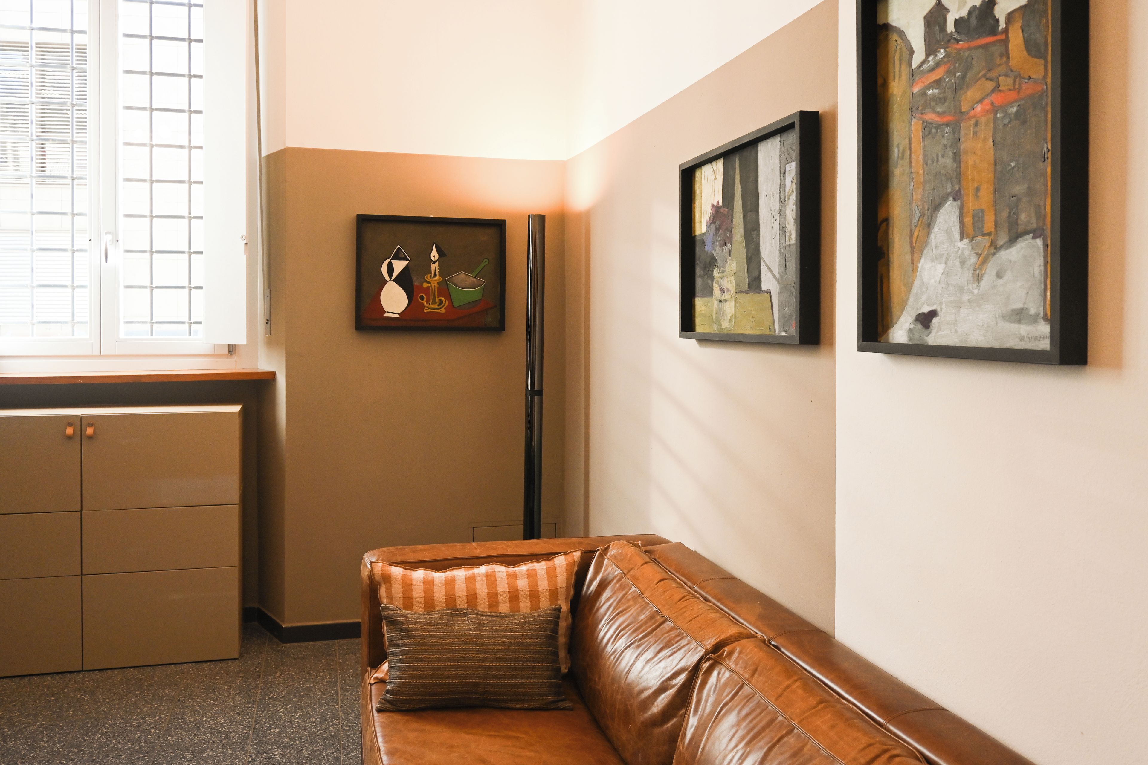 Ein Zimmer mit einem braunen Ledersofa, einem Fenster mit Jalousien und zwei gerahmten Kunstwerken an der Wand.