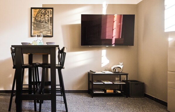Een kamer met een eettafel, stoelen, een aan de muur bevestigde televisie, een klein plankje met boeken en een ingelijste foto aan de wand.