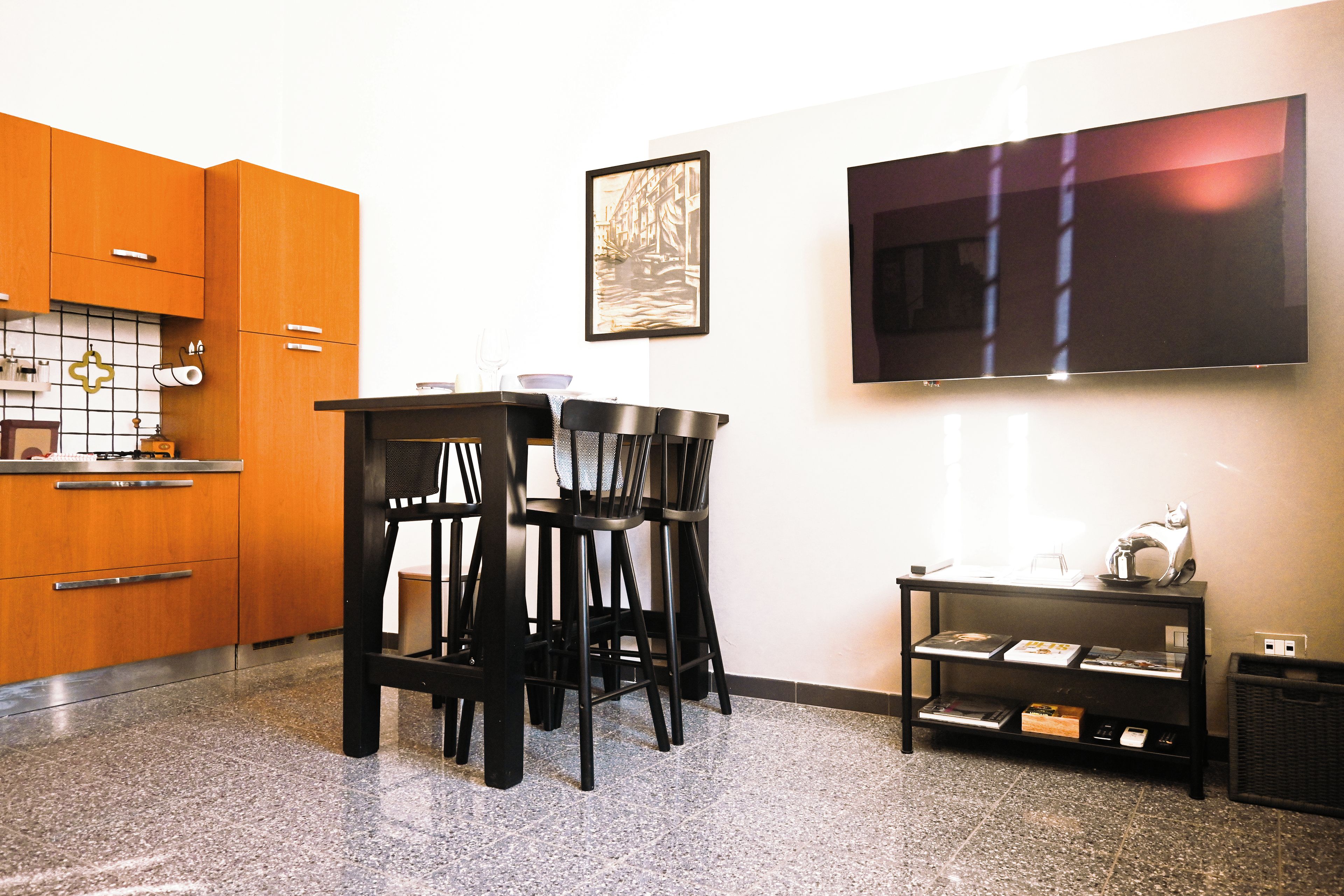 Een moderne keuken met houten kastjes, een eethoek met zwarte stoelen, een flatscreen televisie aan de muur en een kleine tafel met boeken en een katbeeldje.