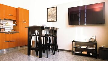 Een moderne keuken met houten kastjes, een eethoek met zwarte stoelen, een flatscreen televisie aan de muur en een kleine tafel met boeken en een katbeeldje.