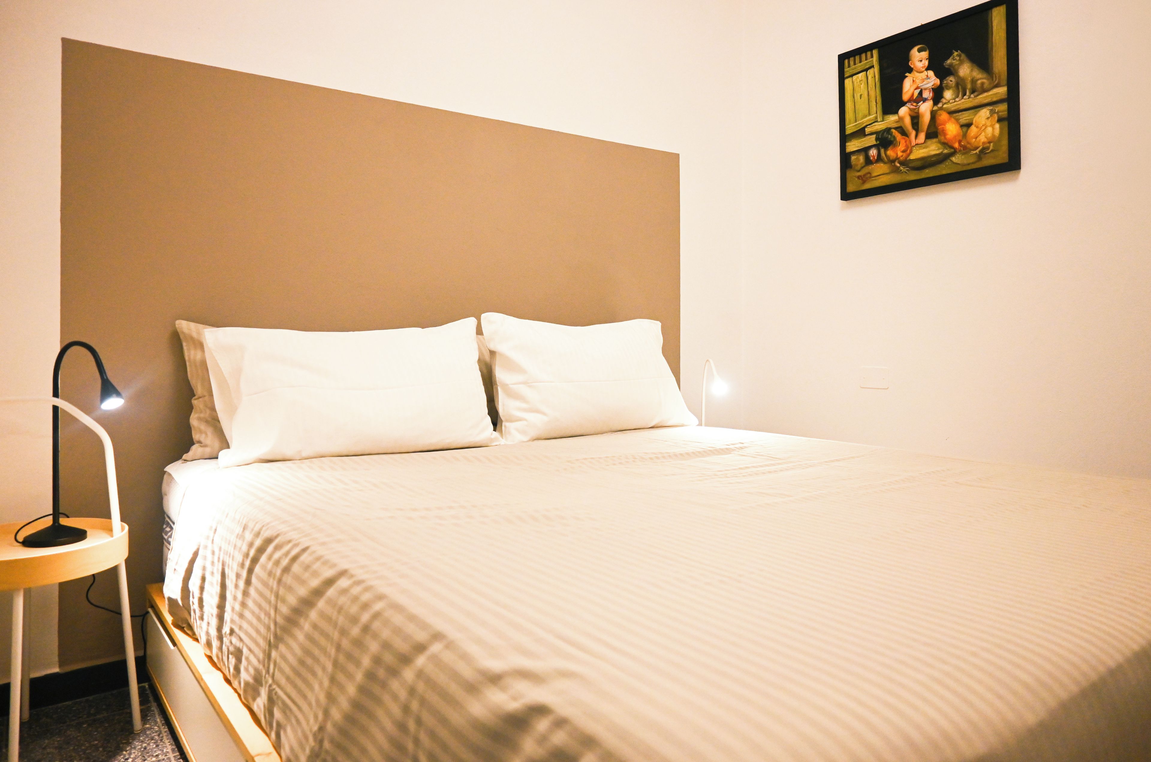 Een bed met witte beddengoed, een nachtkastje met een lamp en een schilderij aan de muur.