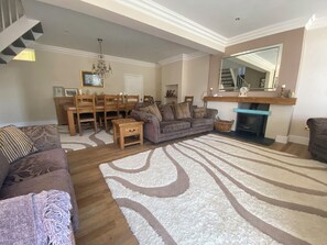 A living room with a fireplace, a dining area with wooden furniture, and a large rug. - Harbour Lodge (Looe)
