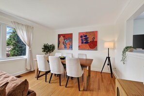 Ein modernes Wohnzimmer mit Essbereich, Holzboden, einer Couch, einem Tisch mit Stühlen, Kunstwerken an der Wand, einer Lampe und Blick durch ein Fenster.