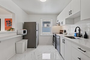 Eine moderne Küche mit weißen Schränken, einem Edelstahl-Kühlschrank, einer Mikrowelle, einem Spülbecken und einem Fenster mit Jalousien.