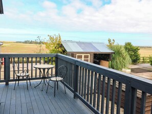 Eine Holzterrasse mit zwei Metallstühlen und einem Tisch, mit Blick auf eine ländliche Landschaft mit einem kleinen Gebäude und Feldern.