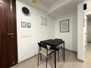 Ein Zimmer mit einer Holztür, einem schwarzen Tisch mit vier Stühlen, einer Wanduhr, einem Korkbrett und einem gerahmten Bild.