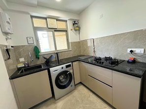 A compact kitchen with a built-in washing machine, a window with blinds, and a countertop with a coffee machine. - Finland II 1BHK_Hirananandani Estate_Spacious (Thane)