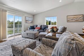 A spacious living room with a large grey sofa, a smaller grey sofa, and a patterned ottoman. There are two windows with views of greenery and a balcony with a railing. - Sturgeon Bay Condo w Marina Slip and in law suite (Sturgeon Bay)