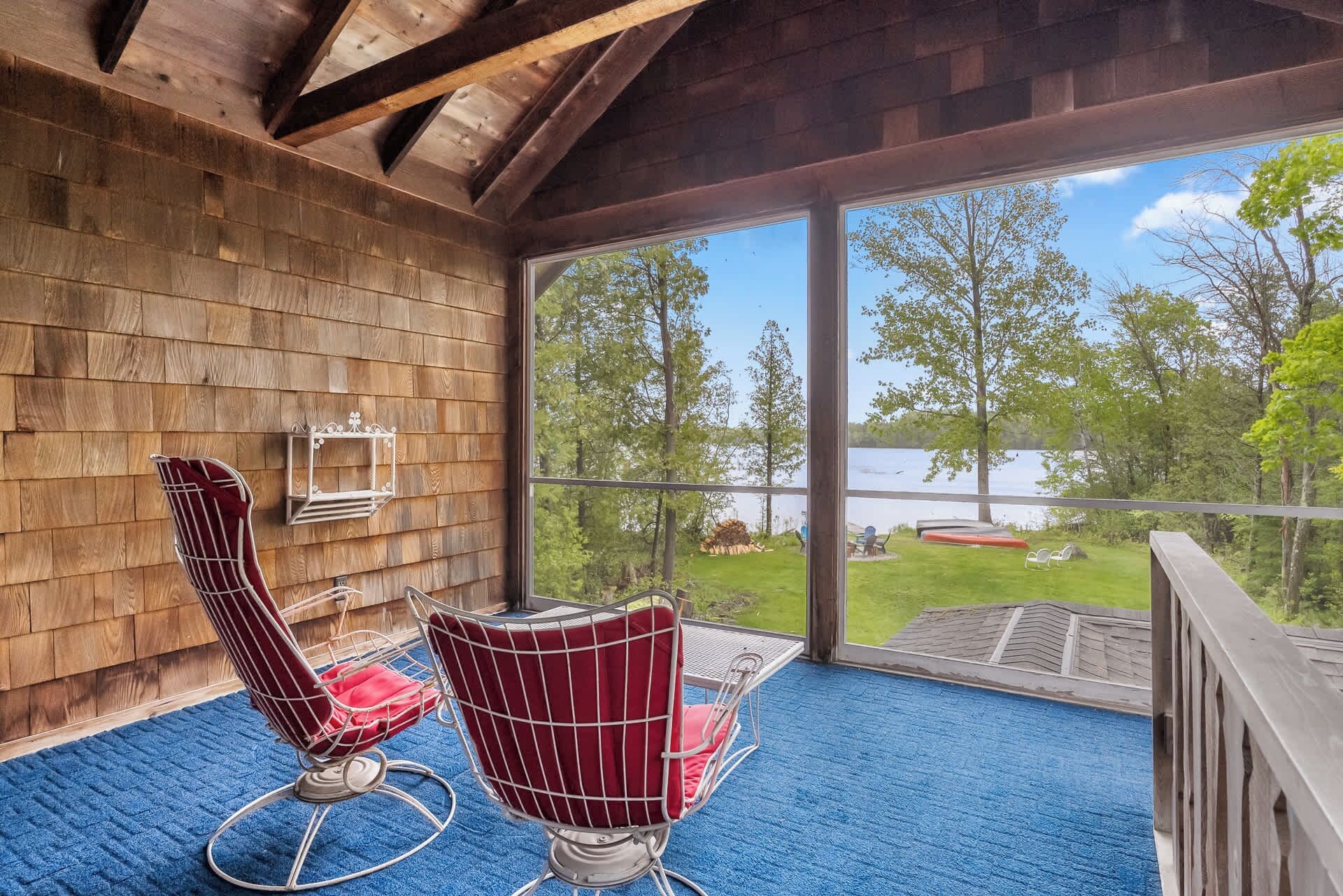 A wooden interior with two red chairs and a table, a large window overlooking a lake and greenery.