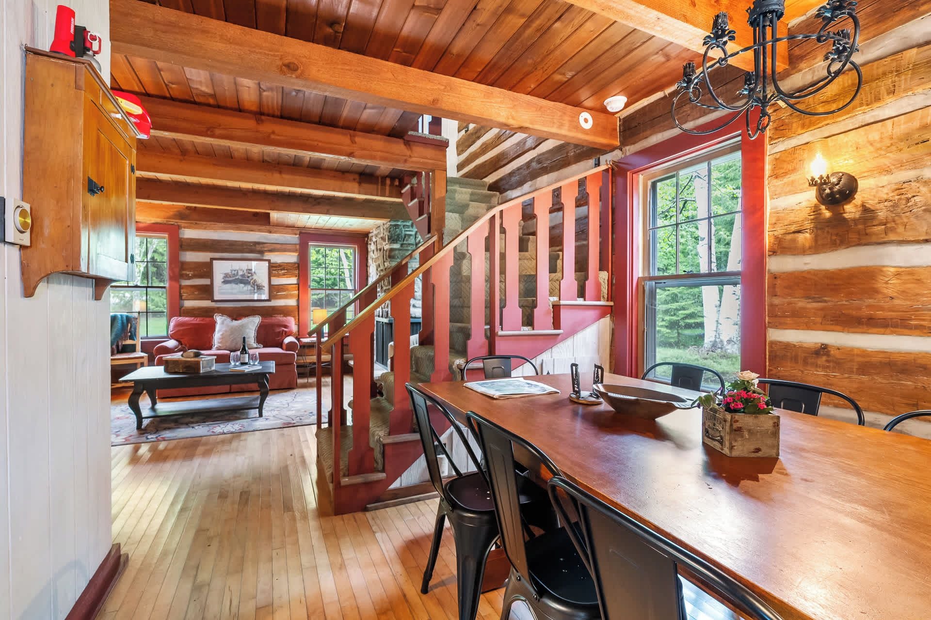 A log cabin interior with a wooden dining table, chairs, a red staircase, and a view of the outdoors.
