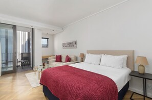 Moderni hotellihuone, jossa on suuri sänky, lasiovi, joka johtaa parvekkeelle, oleskelualue sohvineen ja tuoleineen sekä pieni pöytä.