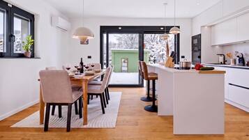 A modern kitchen with a central island, dining area with chairs, and a view through a glass door to a snowy outdoor area.