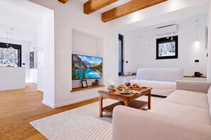Un salon moderne doté d’un téléviseur à écran plat, d’une table basse en bois sur laquelle sont disposés des éléments pour le petit-déjeuner, d’un canapé blanc et d’une grande fenêtre offrant une vue sur la montagne.