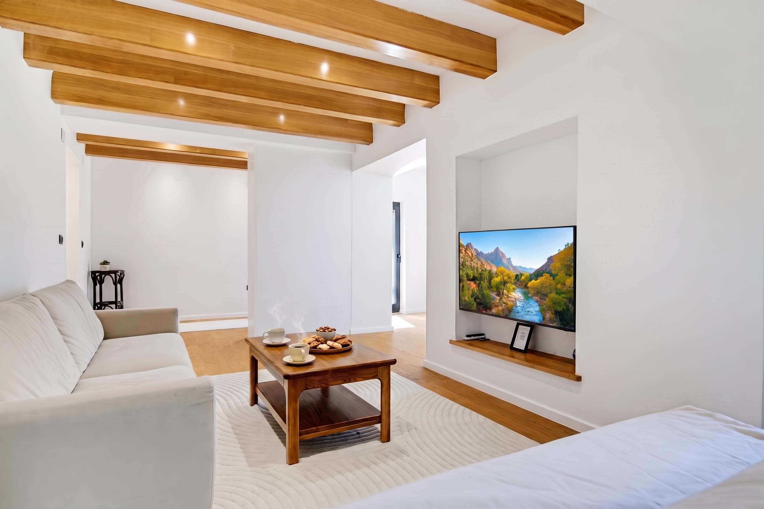 A modern living room with a wooden ceiling, a large flat-screen TV mounted on the wall, a sofa, a coffee table with pastries, and a small side table.