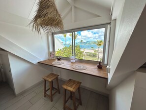 A room with a wooden table and two stools, a window with a view of a beach and palm trees, and a hanging plant. - TI PARADISE Studio SXM Baie Nettlé on the Beach (Marigot)