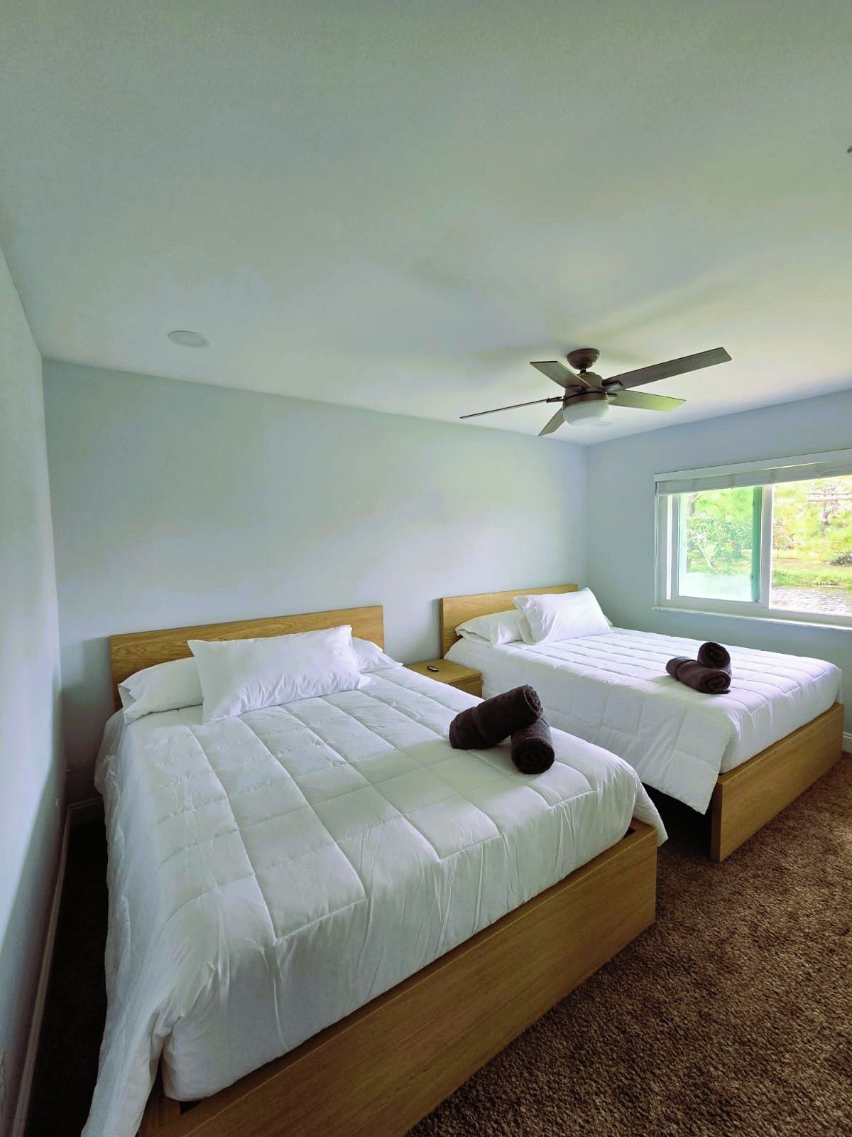 A hotel room with two beds, a ceiling fan, and a window with a view of greenery.