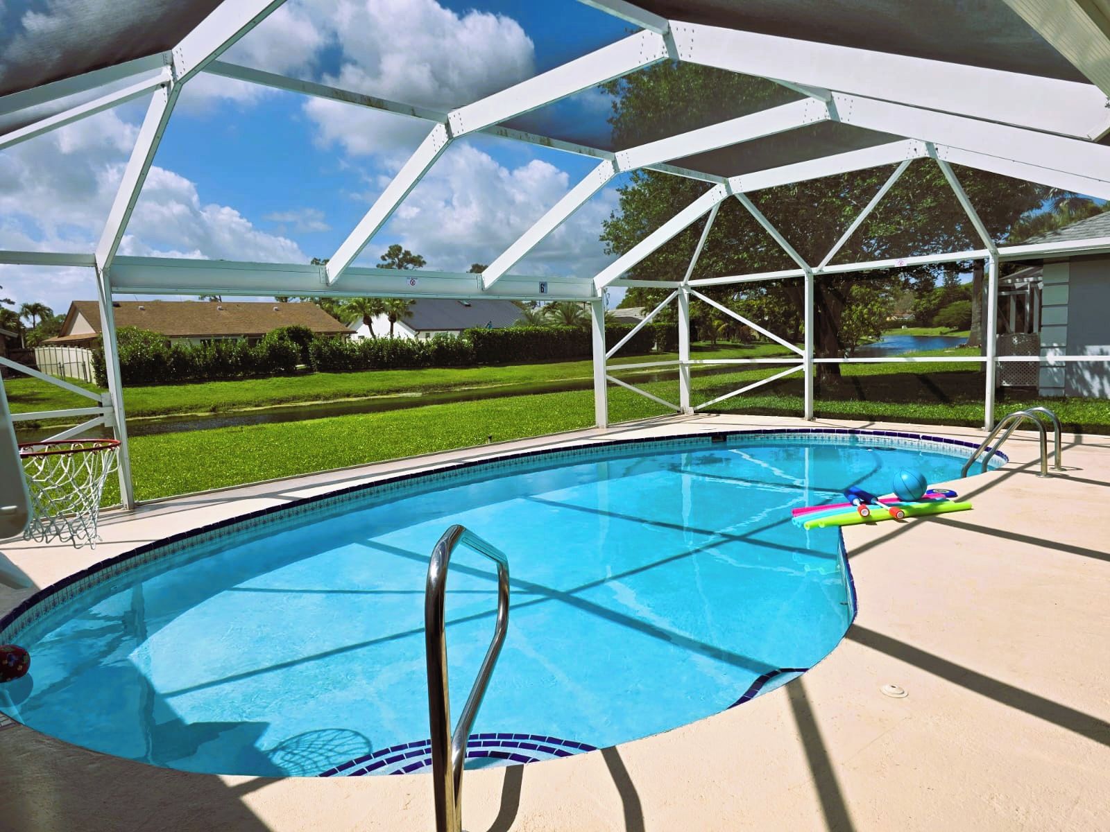 Una piscina con zona techada, canasta de baloncesto y vista despejada de un barrio residencial.