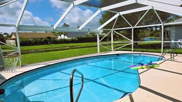 Una piscina con zona techada, canasta de baloncesto y vista despejada de un barrio residencial.