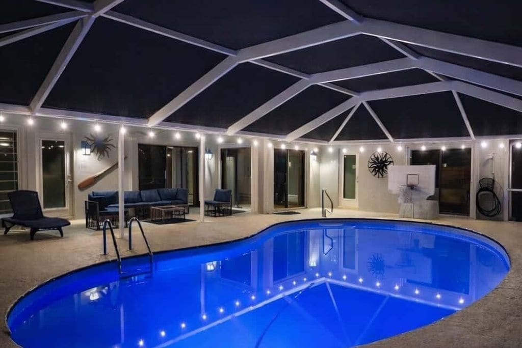 An indoor swimming pool with a glass enclosure, surrounded by a lounge area with seating and a ceiling with a geometric design.