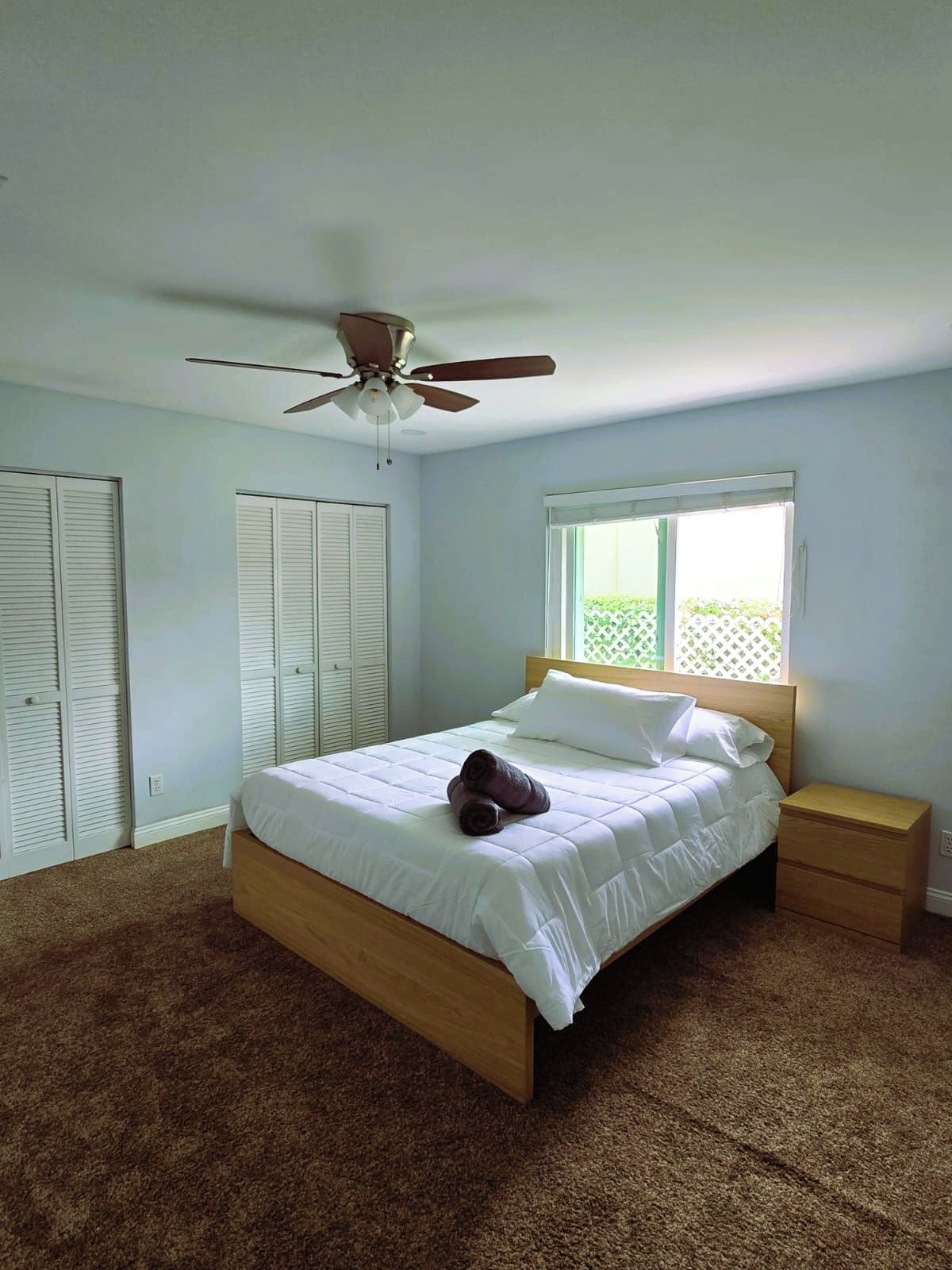 Une chambre à coucher avec un lit, une table de chevet, un ventilateur de plafond et une fenêtre avec des stores.