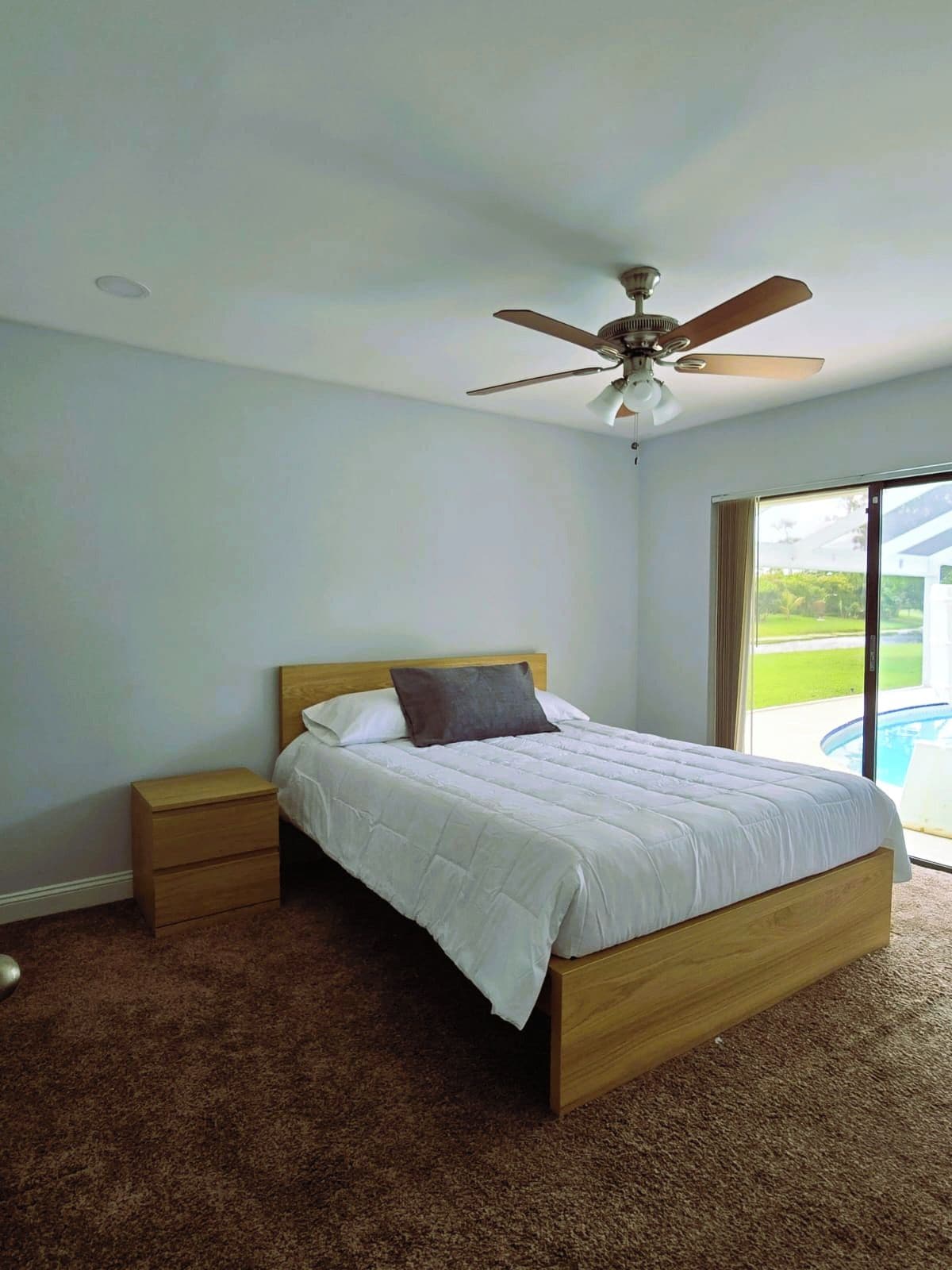 Un dormitorio con cama, mesita de noche, ventilador de techo y vista a una piscina a través de un ventanal amplio.